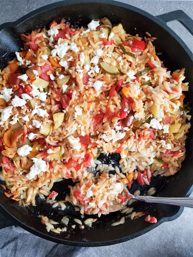 baked orzo with vegetables topped with feta cheese in a cast iron skillet